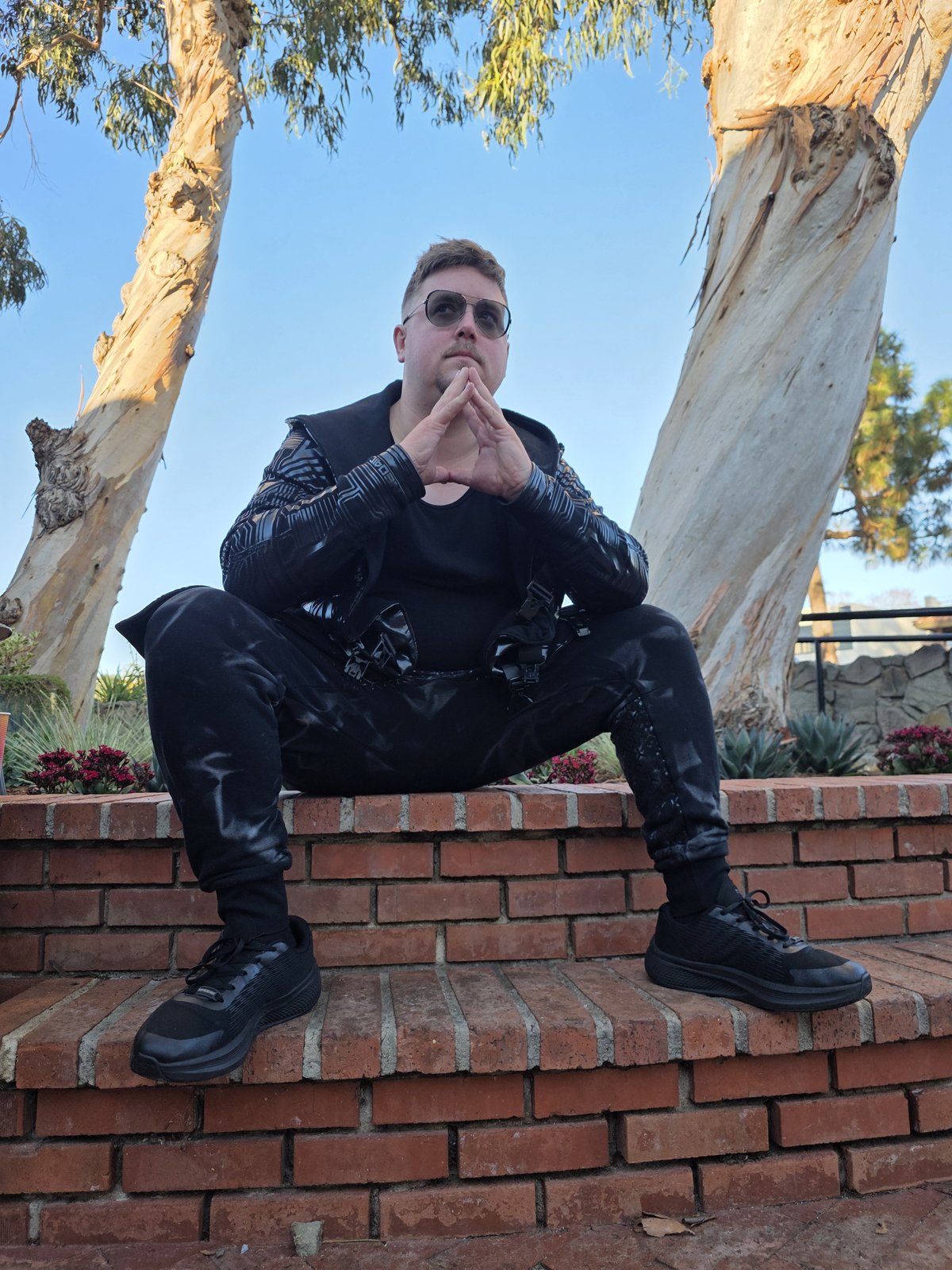 Squirrelybass seated alone on brick steps in black techwear and sunglasses, fingers steepled