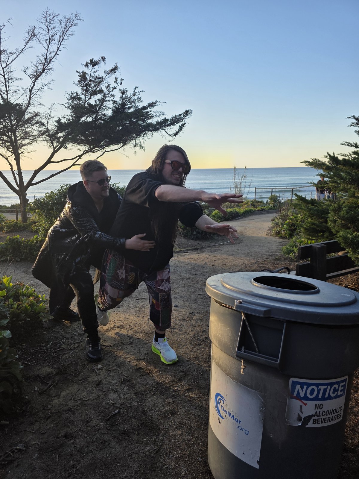Squirrelybass carrying Goose toward a trash can on a coastal path at sunset