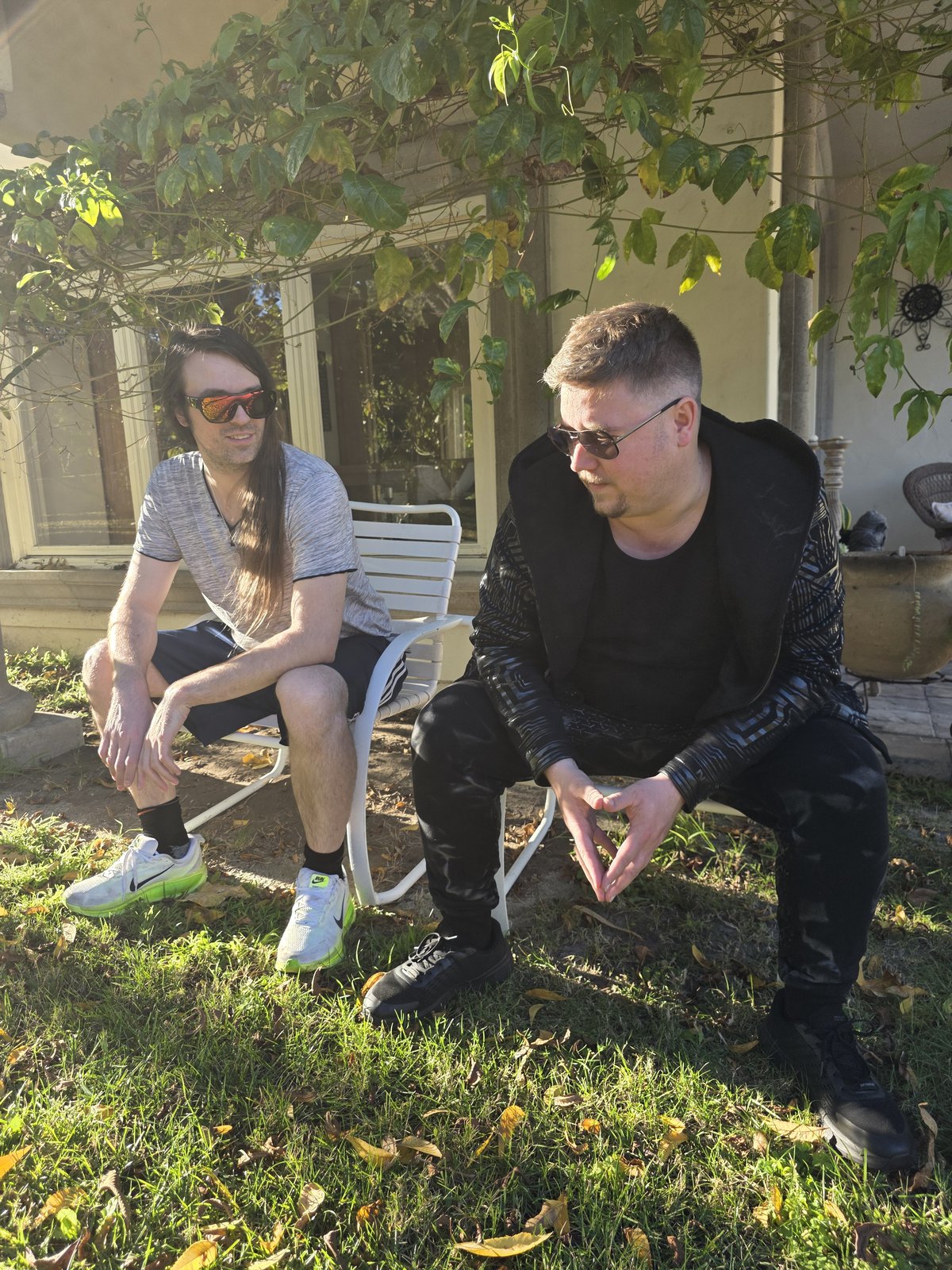 Squirrelybass crouched with steepled hands beside Goose seated on a porch chair under hanging vines
