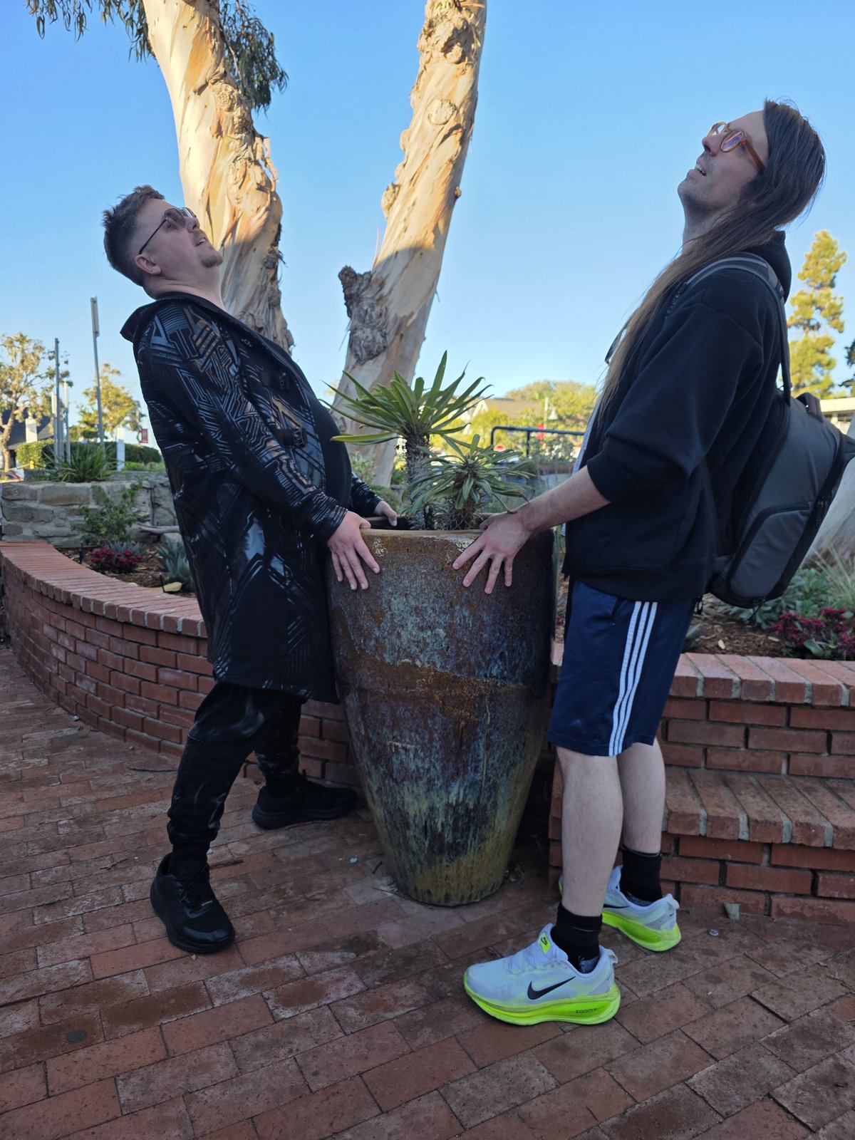 Both artists leaning back dramatically while gripping a large ceramic urn on a brick planter
